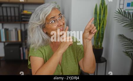 Grey-haired woman pointing and smiling in an indoor office setting with plants and bookshelves in the background, conveying an engaging and welcoming Stock Photo