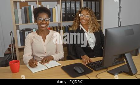 Women working together in an office, with one writing in a notebook and the other working on a computer, both smiling in a professional indoor workpla Stock Photo