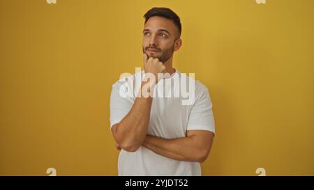 A handsome young hispanic man with a beard poses thoughtfully against an isolated yellow background. Stock Photo