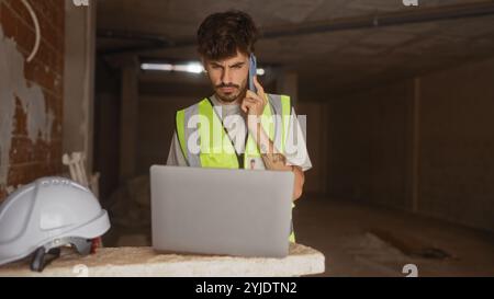Young hispanic man with beard and safety vest working on laptop and talking on phone at construction site Stock Photo