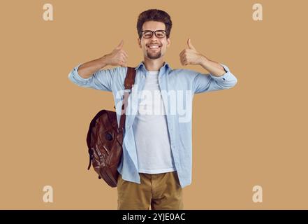 Happy university student guy with leather backpack showing thumbs up on beige background. Stock Photo