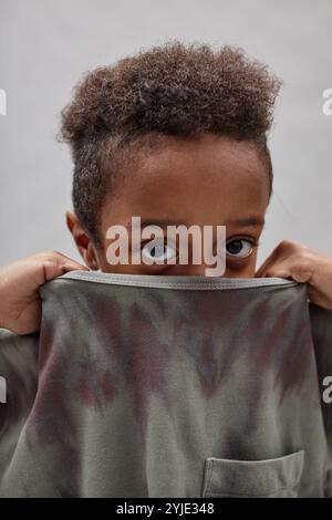 Vertical shot of funny little Black boy hiding face in t shirt pulling up collar and peeking out at camera, while standing against gray background in Stock Photo