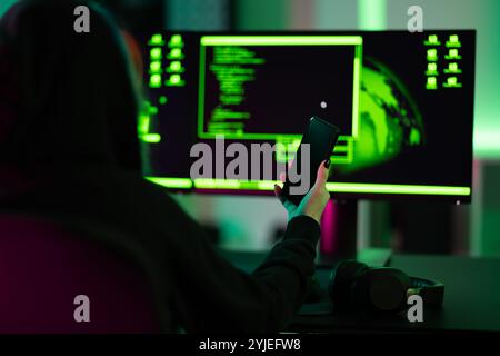 Hacker holding smartphone in front of dual screens with coding interface Stock Photo
