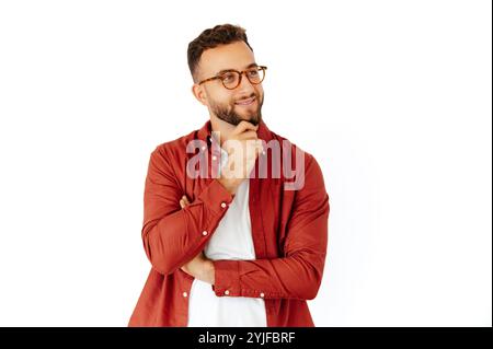 Attractive positive arabian or caucasian young man with glasses, dressed in a red casual shirt with beard dreaming and looking away to the side over white isolated background, smiling Stock Photo
