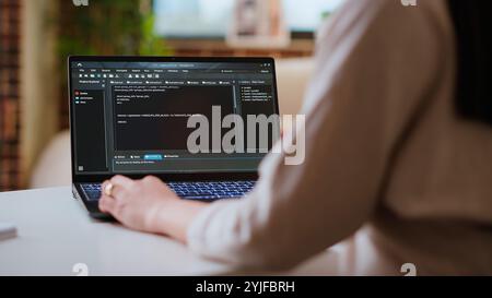 Asian female programmer works on coding software in a home office setup, typing code on a laptop. IT expert uses digital tools for multitasking, software development and app development. Camera B. Stock Photo