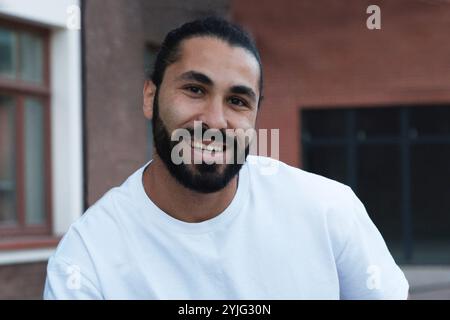 An attractive, laughing dark-skinned man with a beard close-up in a white T-shirt looks at the camera against the background of a house with windows. Stock Photo
