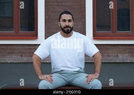 A dark-skinned bearded young man in a white T-shirt and gray trousers sits, looking at the camera, on a bench opposite the facade of a house with wind Stock Photo