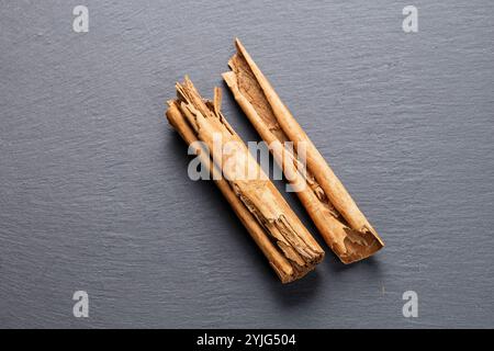 Closeup image of broken pieces of cinnamon sticks scattered on a dark gray slate surface. The cinnamon pieces are light brown, with rough textures and Stock Photo