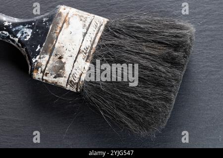 A closeup image of a worn paintbrush with black bristles on a dark slate surface. The brush shows signs of use, with frayed and splayed bristles. Stock Photo