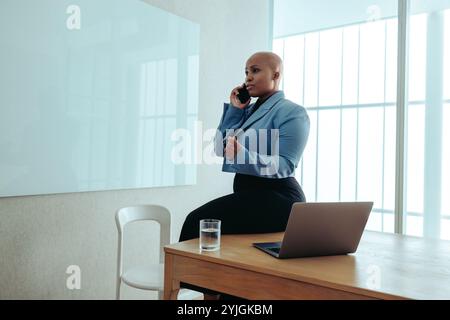 A confident African-American business woman wearing a stylish blue blazer sits on a desk in a contemporary office setting. She engages in a phone call Stock Photo