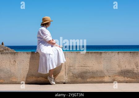 A senior woman sitting on a wall looking at the Mediterranean Sea with a book in her hand, Mallorca Stock Photo