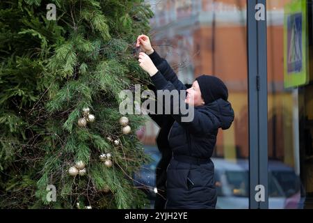 Happy woman in winter clothes decorates Christmas tree on a street, toy balls on fir branches. Preparing to New Year celebration Stock Photo