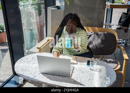 Focused young professional black woman working in a trendy cafe, enjoying a refreshing drink. Urban vibe boosts productivity as she balances work and relaxation stylishly Stock Photo