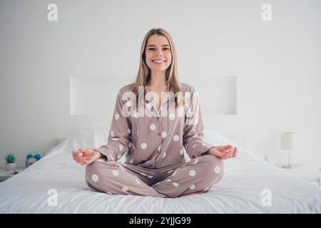 Young woman in polka dot pajamas meditating on cozy bed in bright bedroom during morning light Stock Photo