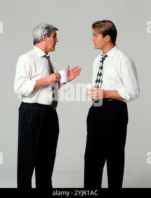 Business & Professions. Two executive men in shirt sleeves, standing indoors with coffee mugs. Stock Photo