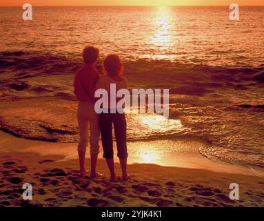 Back view of young couple standing at the beach, watching the sunset. Stock Photo