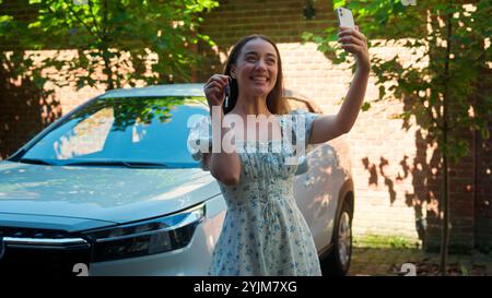 Happy smiling Caucasian woman driver female buyer excited auto owner girl influencer using mobile phone smartphone showing automobile keys talking Stock Photo