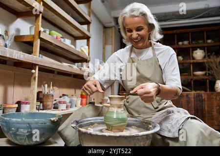 A skilled artisan shapes clay while smiling in her cozy pottery workshop surrounded by tools. Stock Photo