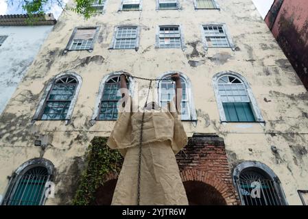 Black woman dressed as a slave, chained and with an iron mask over her mouth and neck, standing with her arms raised against a colonial house. Slavery Stock Photo