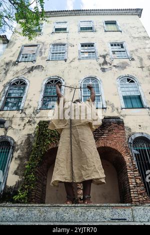 Black woman dressed as a slave, chained and with an iron mask over her mouth and neck, standing with her arms raised against a colonial house. Slavery Stock Photo