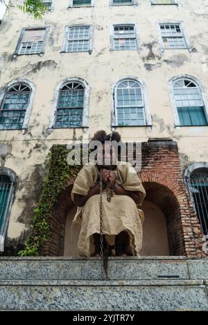 Black woman dressed as a slave, chained and with an iron mask on her mouth and neck, crouched against a colonial house. Slavery in Brazil. Expression Stock Photo