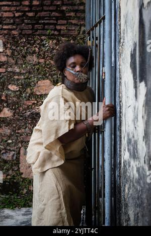 Black woman dressed as a slave, chained with an iron mask on her mouth and neck, holding onto an iron fence. Slavery in Brazil. Representation of the Stock Photo