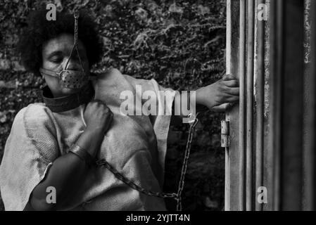 Black woman dressed as a slave, chained with an iron mask on her mouth and neck, holding onto an iron fence. Slavery in Brazil. Representation of the Stock Photo