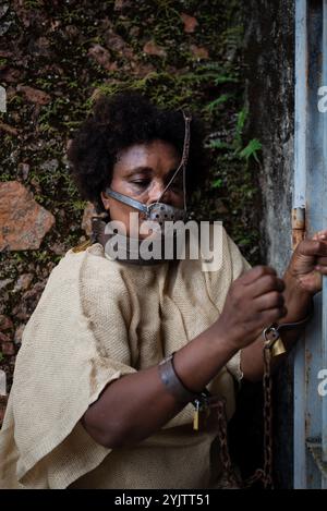 Black woman dressed as a slave, chained with an iron mask on her mouth and neck, holding onto an iron fence. Slavery in Brazil. Representation of the Stock Photo