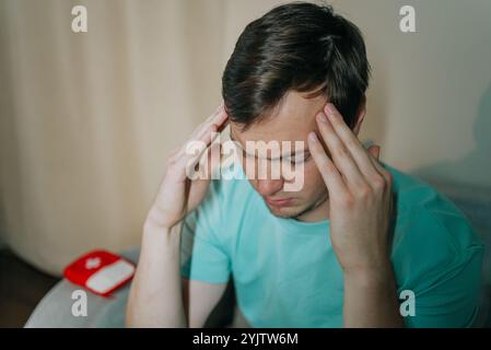 Young man in casual clothes touching his temples, suffering from a strong headache, next to a first aid kit, highlighting the importance of health care for headaches Stock Photo