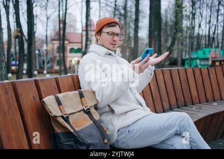 Young man wearing orange winter hat and white jacket is making a video call using his smartphone while sitting on a bench in a city park, expressing confusion and uncertainty Stock Photo