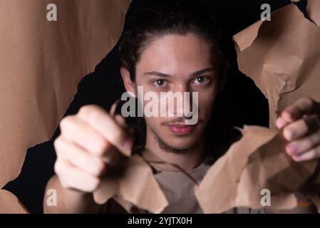 A man coming out through a hole in a sheet of brown paper. Tearing with and showing with his hands the paper. Studio portrait. Stock Photo