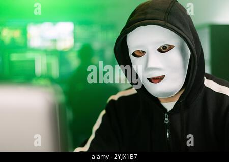 Hacker working on laptop in dark room with green code displayed on monitors Stock Photo