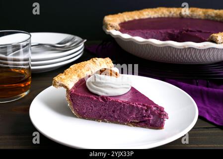 Slice of Purple Sweet Potato Pie Topped with Whipped Cream and a Pecan: Side view of a homemade purple sweet potato pie with a glass of bourbon Stock Photo