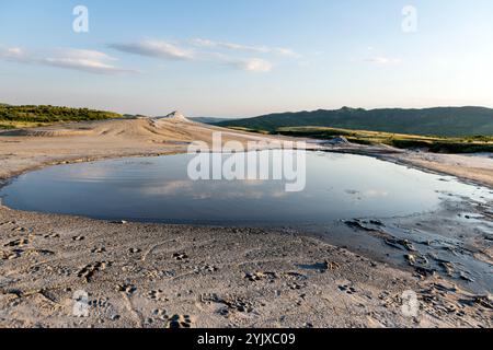 Wide view of a reflective mud pool surrounded by dry, cracked earth with distant hills, showcasing the raw beauty of a mud volcano landscape at sunset Stock Photo