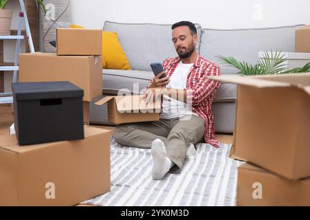 Unpacking boxes on moving day, young man using phone in new home, embracing the spirit of moving. Stock Photo