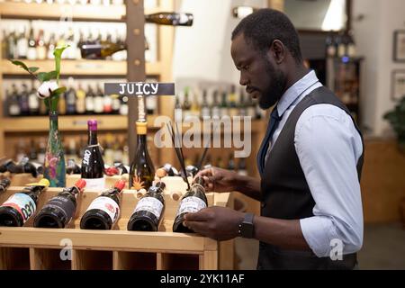 Side view of African American sommelier looking at wine while putting it on wooden rack Stock Photo
