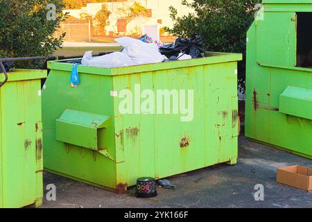 Community dumpsters in a parking lot are overflowing with trash bags while overflowing on the ground around them. Stock Photo