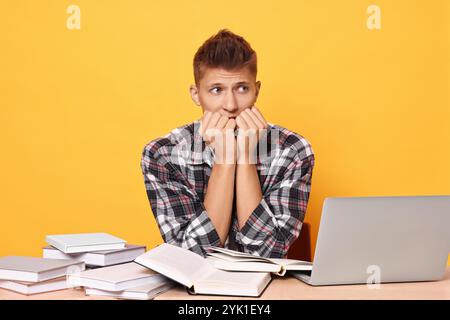 Young student with books and laptop having stress before exam at table against yellow background Stock Photo