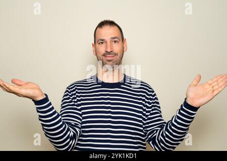 Half length portrait of a bearded hispanic man in his 40s smiling and excited wearing striped sweater and open hands gesture on beige studio Stock Photo
