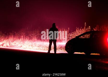 A mysterious hooded figure, silhouetted by car headlights on a country road. With a dark red edit. Stock Photo