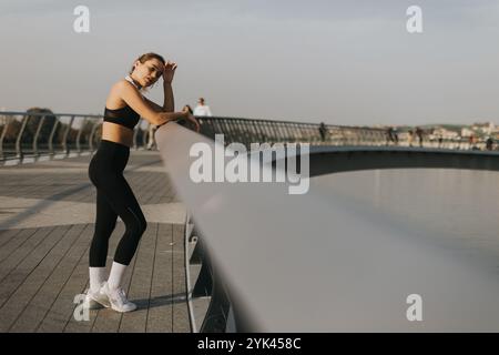 A fit young woman in athletic attire leans on a sleek railing, showcasing her strength and grace while enjoying an evening stroll along the river with Stock Photo