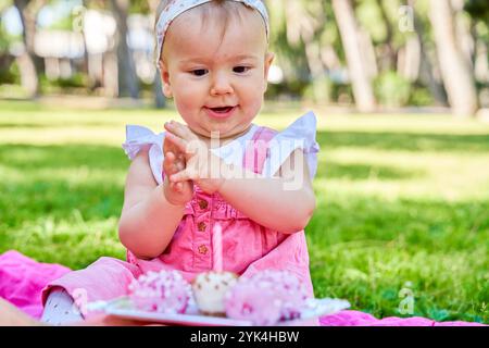 Baby girl in a pink dress clapping with excitement while celebrating her birthday with pink cake pops in a park. Stock Photo