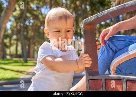 A baby is playing with a bench. The bench is made of wood and has a metal frame. The baby is holding onto the bench with its hands Stock Photo