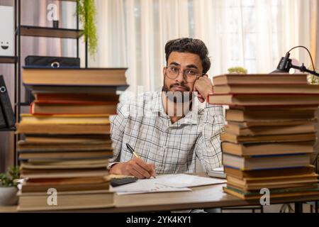 Exhausted Indian businessman reviewing documents reports while using calculator working hard in home office. Stressed tired freelancer touching head in despair surrounded by stacks of books on table. Stock Photo