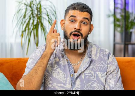 Eureka. Thoughtful clever inspired Indian man make gesture raises finger came up with creative plan feels excited with good idea inspiration motivation, question at home apartment. Guy sits on couch Stock Photo