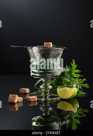 Glass of absinthe with ingredients on a black background. Absinthe spoon, brown sugar, sagebrush branch, and lime slice. Stock Photo