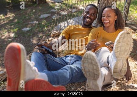 Cheerful African American couple relaxing in a hammock, watching funny videos on a smartphone, enjoying a glamping trip in Europe. Stock Photo