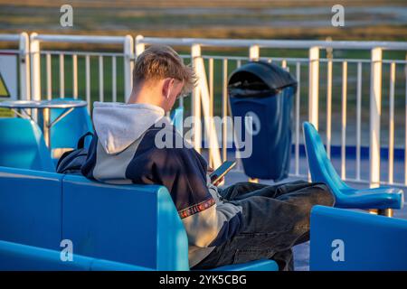 young male or teenage sitting on a bench using a smartphone or mobile phone to communicate or access the internet on the move. Stock Photo