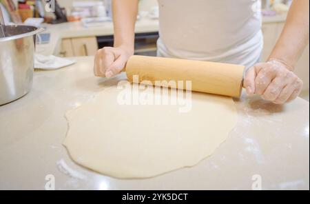 A person is using a rolling pin to flatten dough on a kitchen counter in a cozy kitchen setting, surrounded by baking tools. Stock Photo