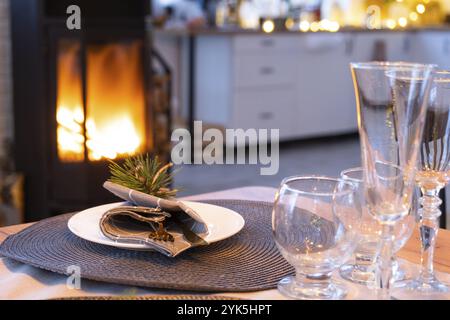 Serving festive table with plates, forks, knives, napkins, glasses close-up near stove fireplace in interior of loft house decorated for Christmas and Stock Photo